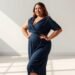 Modelo feminina plus size posando com confiança em ambiente minimalista, vestindo elegante vestido azul marinho com decote em V e manga três quartos. A peça possui modelagem transpassada que valoriza as curvas naturais do corpo, com tecido fluido que proporciona movimento e conforto. A modelo está em posição de três quartos, com uma das mãos apoiada no quadril e expressão sorridente, demonstrando segurança e bem-estar. O cenário apresenta parede neutra em tons claros com jogo de luz natural criando sombras geométricas no chão, proporcionando contraste sofisticado que destaca a silhueta da vestimenta. A iluminação natural realça a textura do tecido e cria uma atmosfera moderna e profissional.